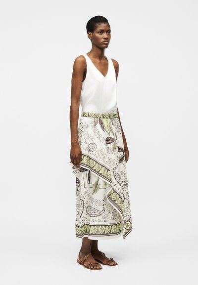 Γυναίκα που φορά άσπρο αμάνικο τοπ με V λαιμόκοψη, συνδυασμένο με ασύμμετρη μίντι φούστα με άσπρο και πράσινο σχέδιο και καφέ φλατ σανδάλια.