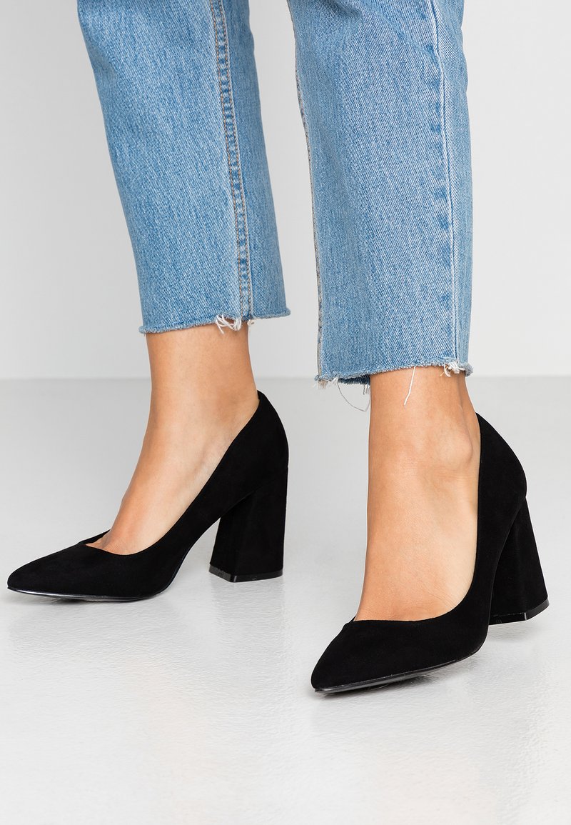 Black suede high-heeled pumps with a pointed toe and chunkier heel, paired with cropped blue denim jeans featuring frayed hems.