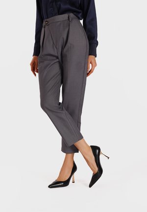 Pantaloni grigi su misura con vita alta, pieghe frontali e un dettaglio con bottone, abbinati a décolleté nere a punta e una camicetta blu scuro.