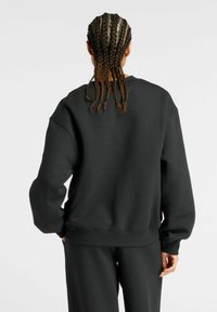 Sweatshirt noir à coupe décontractée, fabriqué en tissu épais, avec des épaules tombantes et des poignets côtelés. Porté avec un pantalon assorti.
