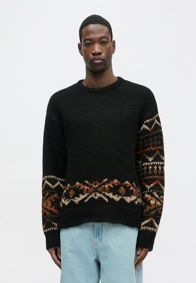 Fekete kötött pulóver földszínekkel díszített mintákkal. Kereknyakú és bordázott mandzsettákkal rendelkezik. Világoskék farmernadrággal viselve.