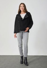 Mustast fliisist lukuga jope kõrge kraega, mis on kombineeritud helehalli teksade ja mustade pahkluu saabastega, neutraalse taustaga.