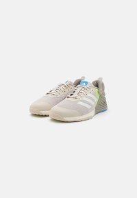 Light beige athletic shoes with mesh upper, textured accents, blue logo detail, and a green-tinted sole. Features rounded toe and laces.