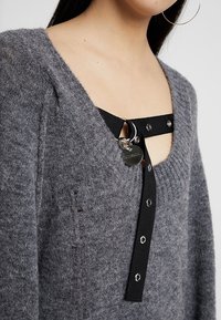 Grauer Strickpullover mit V-Ausschnitt, ausgestattet mit schwarzen Trägern und einem silbernen Ringakzent. Texturiertes Material mit sichtbaren Nähdetails.