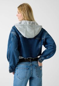 Przycięta kurtka dżinsowa z szarą kapturem, kontrastującymi odcieniami niebieskiego, postrzępionym dołem i oversize'owymi rękawami. Noszona z jasnoniebieskimi dżinsami i czarnym paskiem.