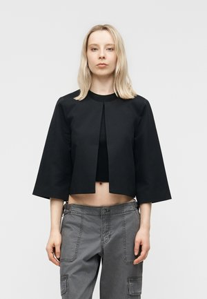 Vrouw met lichtblond haar staat voorover gericht en draagt een zwarte cropped jas, een zwart topje en grijze cargobroek tegen een effen lichte achtergrond.