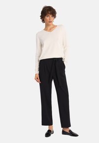 Sweter w kolorze kremowym, dziany w serek, zestawiony z czarnymi spodniami o wysokim stanie z wiązanym detalem w talii. Modelka w czarnych mokasynach.