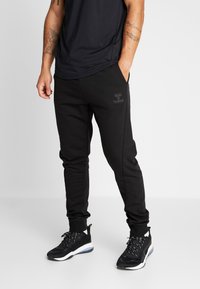 Svarta träningsbyxor med slim fit, elastisk midja, sidofickor och en diskret logotyp på låret. Matchas med svarta sportskor.