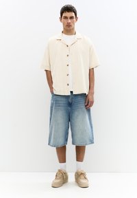 Light beige short-sleeve shirt with wooden buttons, paired with blue denim shorts. Beige sneakers and white socks complete the casual look.