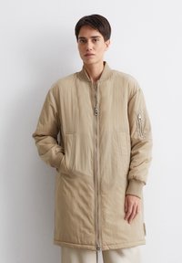 Beige bomberjack met een ritssluiting aan de voorkant, ribgebreide manchetten en twee grote zijzakken. Glad, lichtgewicht stof met een minimale textuur.