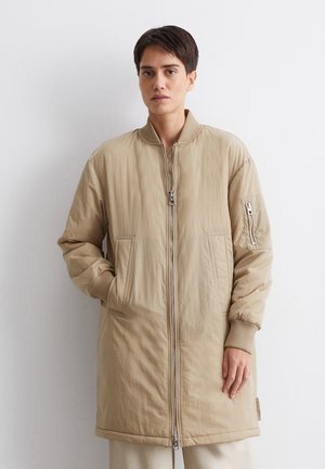 Beige bomberjack met een ritssluiting aan de voorkant, ribgebreide manchetten en twee grote zijzakken. Glad, lichtgewicht stof met een minimale textuur.