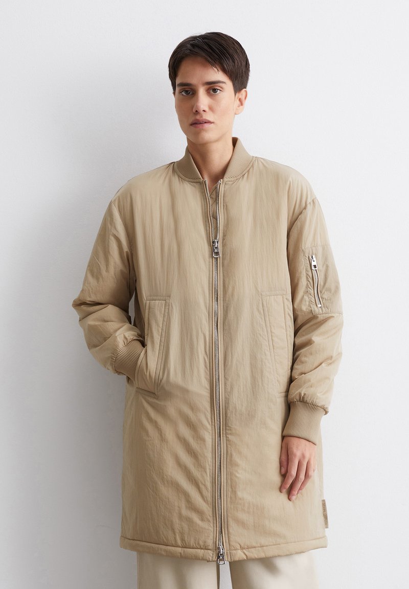 Beige bomberjack met een ritssluiting aan de voorkant, ribgebreide manchetten en twee grote zijzakken. Glad, lichtgewicht stof met een minimale textuur.