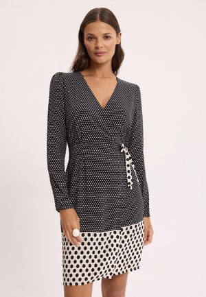 Kobieta ubrana w czarną, długorękawową sukienkę kopertową z białymi kropkami na górze i większymi czarnymi kropkami na białym brzegu spódnicy.