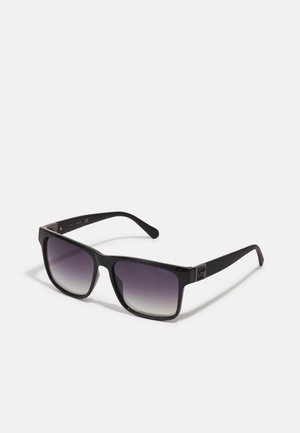 Occhiali da sole con montatura in plastica nera, lenti quadrate con un gradiente che va dal grigio scuro al chiaro. Dettagli in metallo sulle astine.