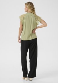 Blusa verde chiaro con maniche corte in pizzo, abbinata a pantaloni neri larghi. La blusa ha un colletto alto e una texture liscia.