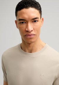 Beige t-shirt met ronde hals van zachte stof, met korte mouwen en een delicaat geborduurd logo op de borst. Zachte textuur.