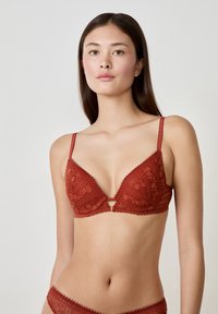 Bralette en dentelle couleur orange brûlé avec une forme triangulaire, des bretelles réglables et un détail en forme de trou de serrure au centre. Finition à bords festonnés.