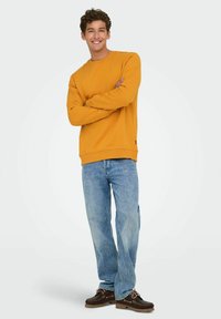 Geel sweatshirt gemaakt van zachte stof, gecombineerd met lichtblauwe, losse jeans en donkerbruine schoenen met veters. Model staat met gekruiste armen.