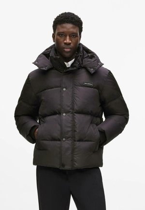 Mężczyzna w czarnej kurtce puchowej z wysokim kołnierzem i kapturem, ręce w kieszeniach, neutralne tło.