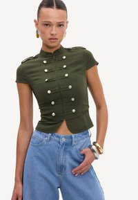 Jeune femme portant un haut ajusté à manches courtes style militaire vert olive avec des boutons argentés et un jean taille haute bleu clair.