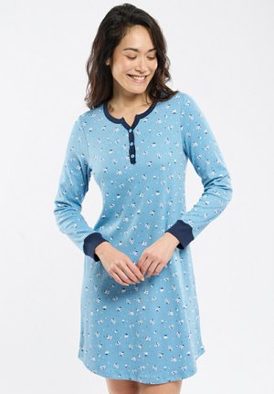 Femme souriante, regardant vers le bas, portant une robe à manches longues bleue avec de petits motifs floraux blancs et un col et des poignets bleu foncé.