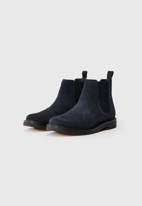 Une paire de bottes Chelsea en suède bleu marine foncé avec des panneaux élastiques noirs sur les côtés et des languettes de tirage sur un fond blanc.