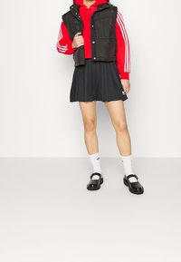 Gilet imbottito nero sopra una felpa rossa corta, abbinato a una gonna plissettata nera. Calze bianche con logo e scarpe Mary Jane nere.