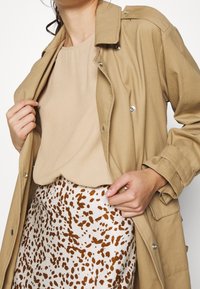 Ljusbeige trenchcoat med silverfärgade knappar, lager över en beige topp och en prickig krämfärgad och brun mönstrad kjol.