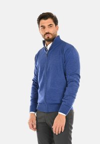 Maglione blu con zip realizzato in tessuto lavorato a maglia, con colletto alto, polsini a costine e dettagli con zip, indossato sopra una camicia bianca.