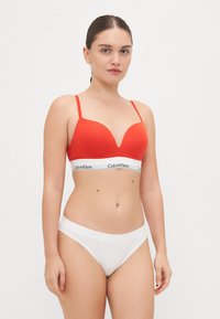 Crvena braletica s dubokim izrezom, tankim naramenicama i pojasom s Calvin Klein logom, uparen s bijelim hipster gaćicama koje imaju glatku teksturu.