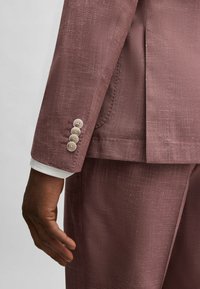 Ljus burgundyfärgad kavaj i texturerat tyg. Har fyra krämfärgade knappar vid ärmen och en vit skjortkrage under ärmen.