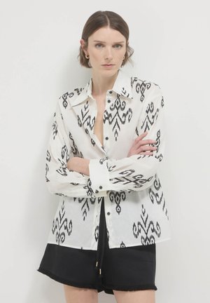 Weißes Hemd mit schwarzem Ikat-Muster, langen Ärmeln, Kragen und schwarzen Knöpfen; kombiniert mit schwarzen Shorts.
