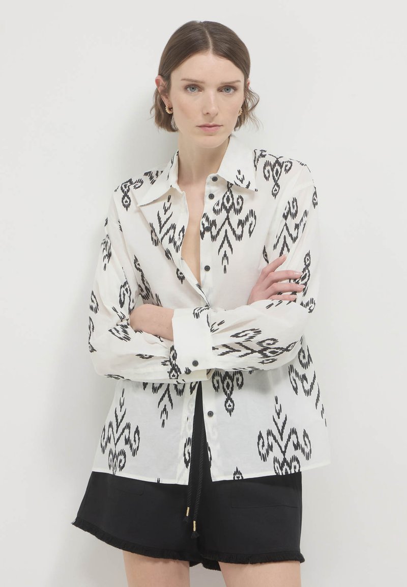 Camicia bianca con motivo ikat nero, maniche lunghe, colletto e bottoni neri; abbinata a pantaloni corti neri.