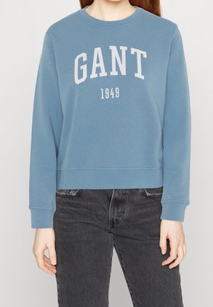 Persoon die een blauwe GANT-trui draagt met de tekst "GANT 1949" en een zwarte spijkerbroek, getoond van schouders tot heupen.