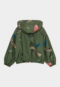 Giacca a vento verde con vestibilità ampia, caratterizzata da varie etichette e timbri stampati in rosso, blu e beige. Vita e polsini elasticizzati.