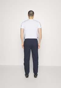 Enges weißes Sportshirt mit kurzen Ärmeln, kombiniert mit dunkelblauen Jogginghosen. Schwarze Sportschuhe mit orangefarbenen Akzenten vervollständigen das Outfit.