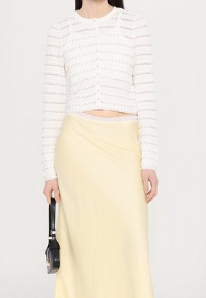 Mujer que lleva una blusa blanca de manga larga abotonada con detalles de perlas y una falda amarillo pálido, sosteniendo un pequeño bolso negro en su mano izquierda.
