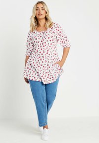 Blusa blanca con patrón floral rojo, mangas abullonadas largas, cuello con cordón y bolsillos, combinada con jeans de mezclilla azul claro y zapatillas blancas.
