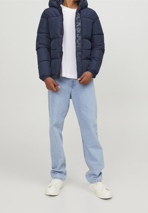 Doudoune marine avec capuche, fermeture éclair avant, texture douce et logo en relief. Associée à un jean bleu clair et des baskets blanches.