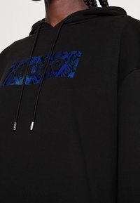 Felpa nera con un logo a patch a serpente blu con il testo "HUGO." Dettagli con cordini e punte metalliche argentate. Texture morbida.