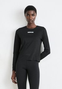 Vrouw in een zwarte lange mouwen top van Calvin Klein en zwarte leggings staat tegen een plain witte achtergrond, en kijkt recht in de camera.
