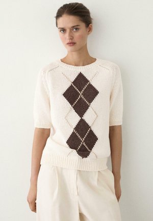 Žena v krémovém svetru s krátkými rukávy a hnědým vzorem argyle, v kombinaci s krémovými kalhotami s vysokým pasem, stojící před bílou zdí.