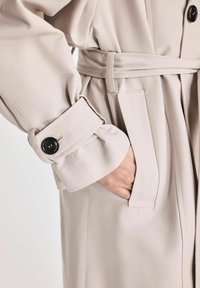 Beżowy płaszcz trench z paskiem, z szerokimi rękawami, kieszenią oraz czarnym guzikiem na mankiecie. Gładka, strukturalna tekstura materiału.