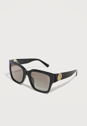 Black square-frame sunglasses with gradient lenses and gold circular logo accents on the temples, set against a plain light background.