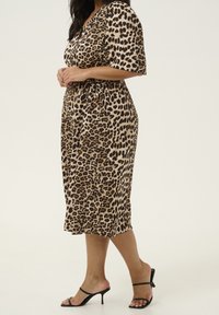 Frau in einem midi Kleid mit Leopardenmuster und Bindegürtel, kombiniert mit schwarzen Absatzsandalen, steht vor einem schlichten hellen Hintergrund.