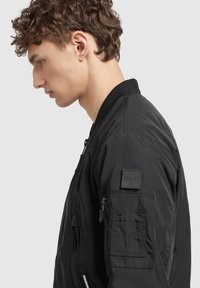 Schwarze Bomberjacke aus glattem Stoff, mit einem durchgehenden Reißverschluss, geripptem Kragen und einer Tasche am Ärmel mit einem Markenetikett.