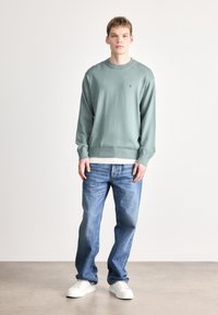 Πράσινο πουλόβερ με μακριά μανίκια και στρογγυλή λαιμόκοψη, συνδυασμένο με ανοιχτό μπλε τζιν παντελόνι κανονικής γραμμής και λευκά αθλητικά παπούτσια. Λεπτομέρεια με μινιμαλιστικό λογότυπο.