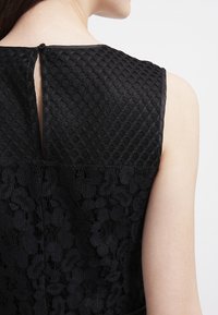 Svart klänning med en texturerad bodice med diamantmönster och en spetstredel nedtill. En nyckelhålsrygg med en knapppdetail syns.