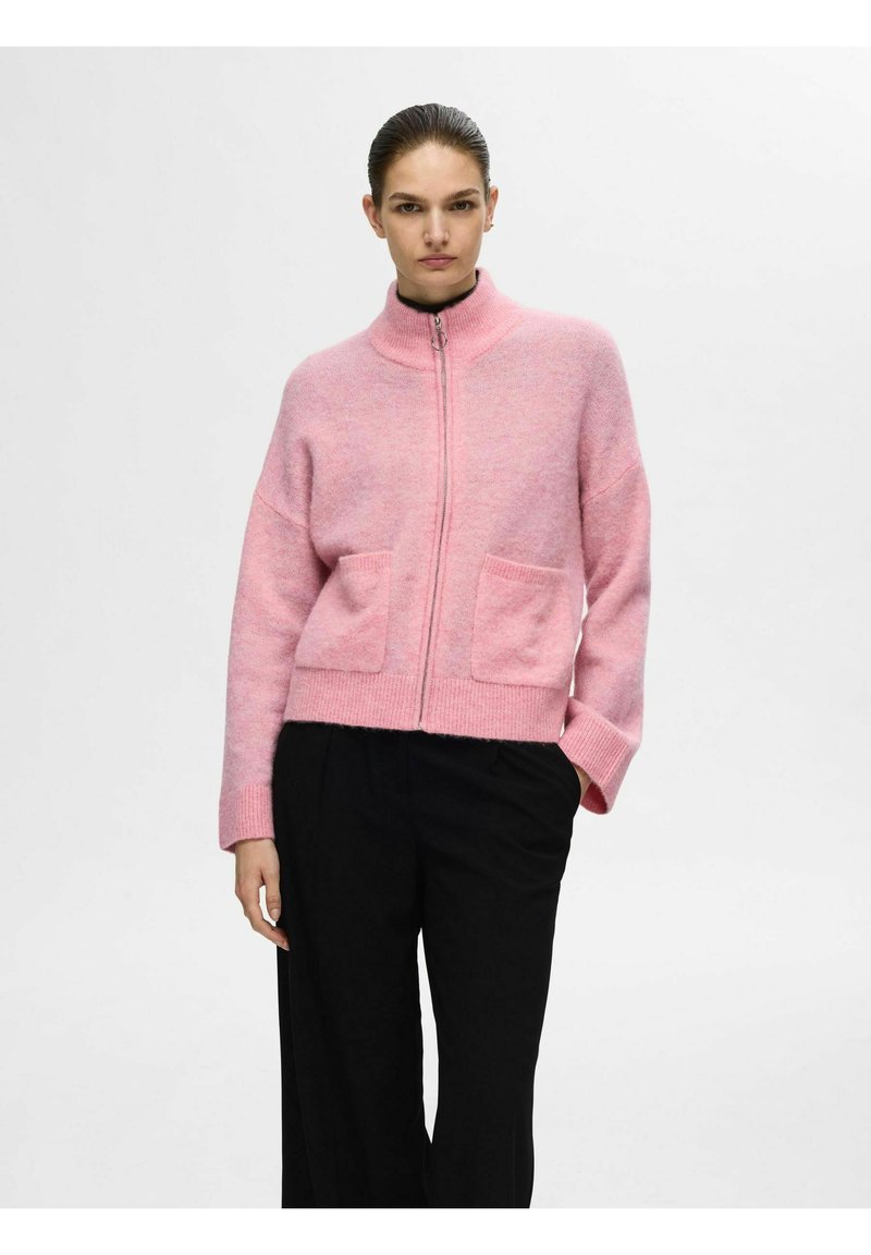 Cardigan zippé rose avec col haut, deux poches avant et poignets côtelés. Sa texture tricotée lui confère un aspect doux. Porté avec un pantalon noir.
