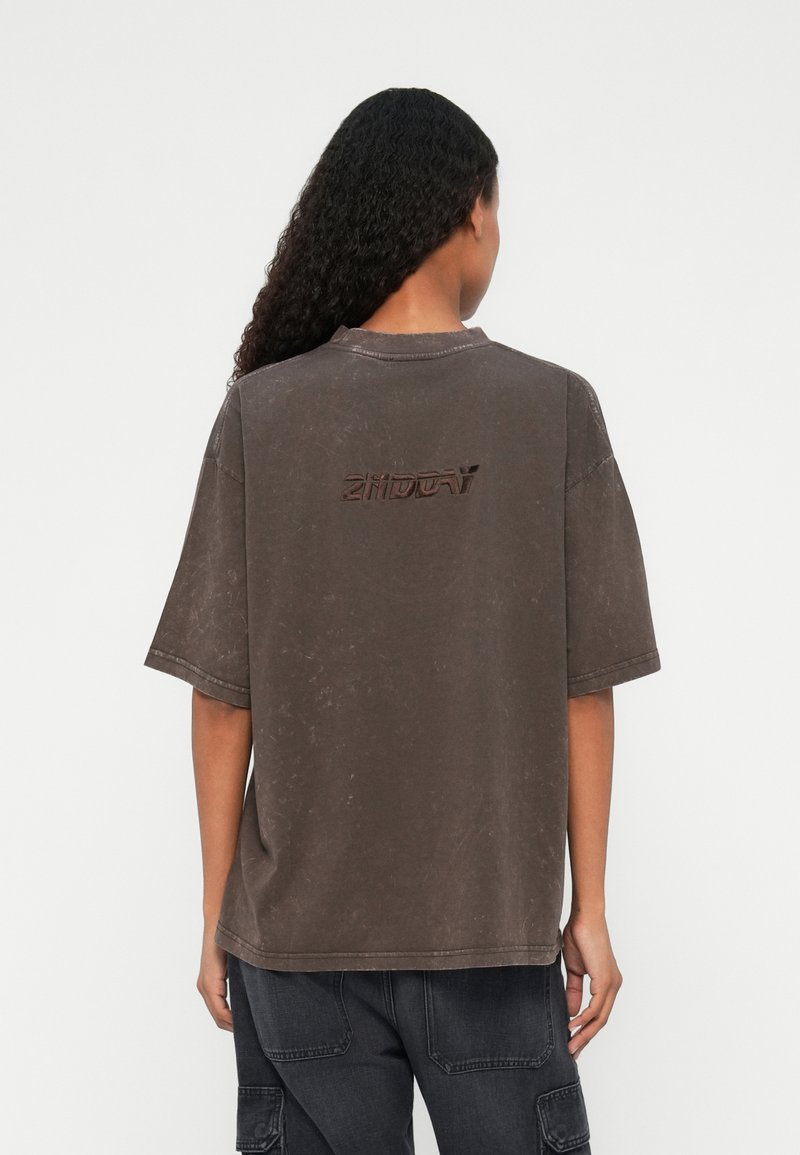 Person med langt krøllet hår iført en løs, falmet mørkebrun T-shirt med tekst på ryggen og mørke cargo-bukser mod en hvid baggrund.